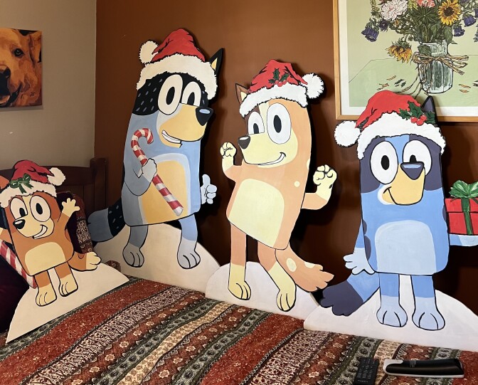 Cut outs of Bluey characters in Christmas hats placed on a bed with a printed sheet. A photo of a dog and a picture of a flower vase are visible. 