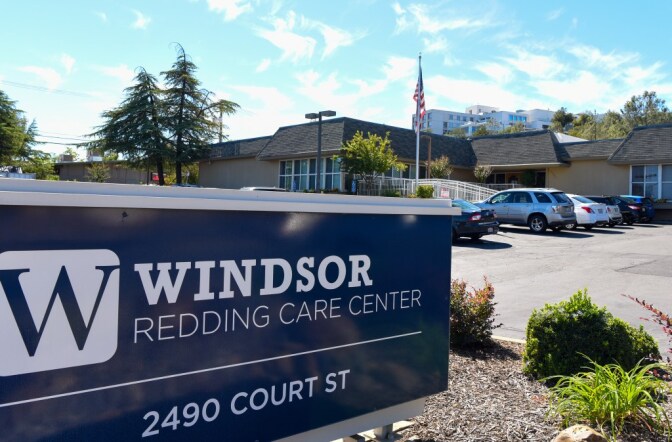 A sign is shown outside a nursing home facility. The sign is navy and rectangular, positioned horizontally, and reads "Windsor Redding Care Center 2400 Court St" in capital white letters. The facility is shown behind the sign, a one-story beige building with an American flag flying out front and cars in the parking lot. 