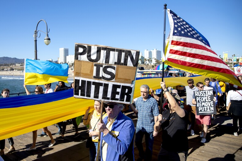 A crowd of one dozen people walk toward the end of the Santa Monica pier with buildings, the beach, the ocean and shore in the background. A long Ukrainian flag colored blue and gold is stretched along the entire length of the crowd along the center. At the front of the crowd, and a gray-haired man with dark sunglasses, a blue jacket holds a brown cardboard sign with letters in black outlined in white that reads "PUTIN IS HITLER." A man wearing a dark shirt next to him holds a flag pole bearing two flags, the American flag, with red and white stripes, a blue rectangle and white stars, and a Ukrainian flag, blue on top and gold on the bottom. Behind that man is a young man in his twenties wearing pink shorts and a blue shirt holding a black sign with white lettering that says "Russian People Remove Putin."