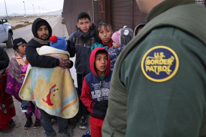 A family who appear to be seeking asylum stand in front of an American border patrol agent. Both the mother and father are carrying babies, and a young boy with a hood looks up at the agent with a sad but hopeful facial expression. They family appears weary. 