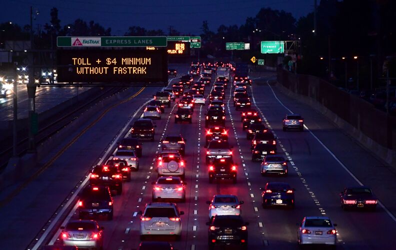 Traffic on a five-lane highway in Los Angeles is shown from behind in an aerial view, so that brake lights are shown. It is night time. A digital sign on the left of the photo reads "TOLL + $4 MINIMUM WITHOUT FASTRAK" in yellow capital letters on a black background.