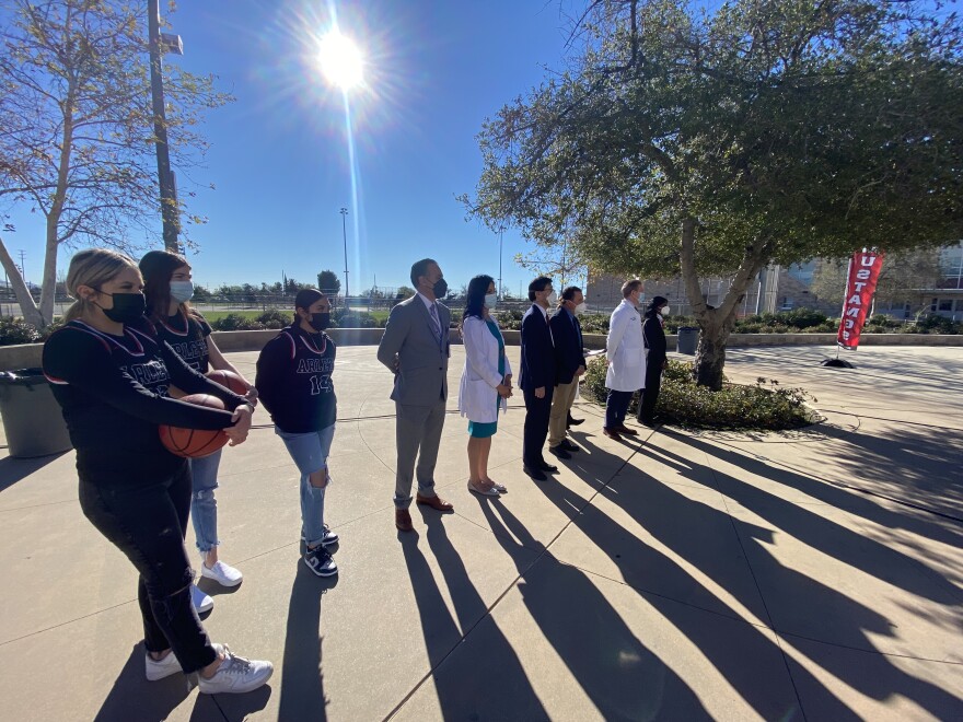 In the courtyard of a high school, a row of people stand in a line with the sun at their backs, with the sunlight casting long shadows onto the pavement. Two younger women at left hold basketballs at their stomachs. Others in the line are wearing business attire, and two people wear white doctor's coats.