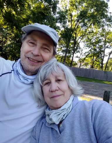 Two people with light skin tone smile into the camera for a selfie together. One, a man, is wearing a light-colored hat and sweater, with what looks like a light grey turtleneck underneath. The other, a woman, is wearing a grey sweaters with a white scarf.