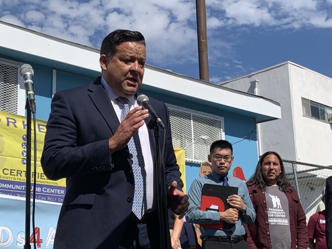 A middle-aged Latino in a dark suit and tie speaks outdoors in front of microphone, while two men stand to his left.