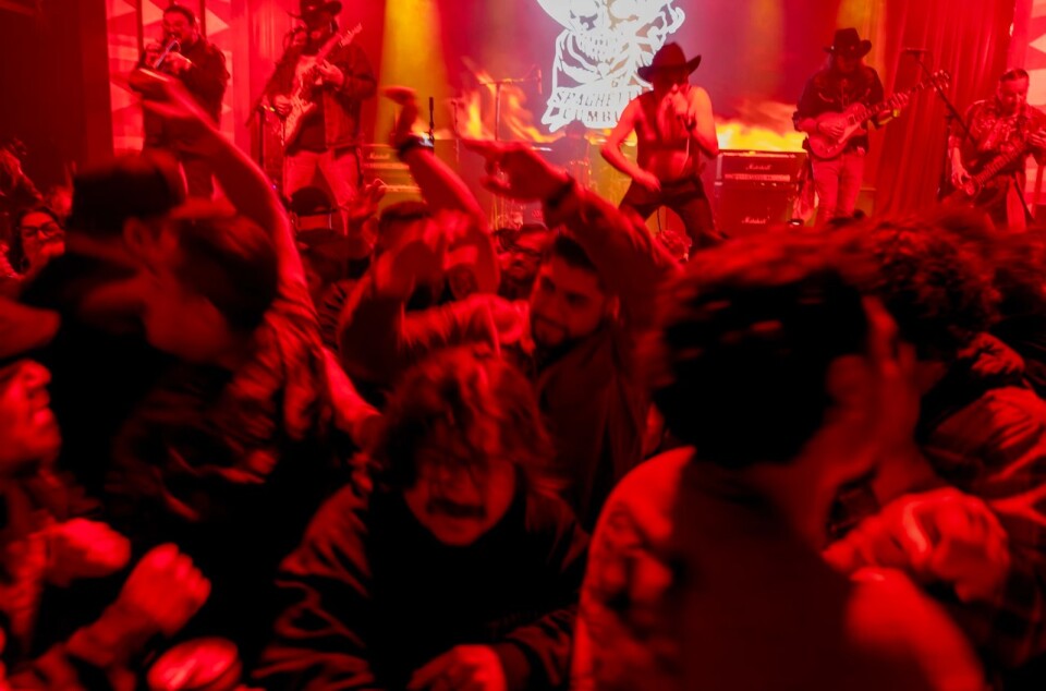 A crowd of young men dance together in front of a stage where a band of men with instruments perform.