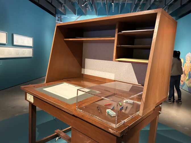 A dark wooden drawing desk on a blue-green platform in the middle of a room. Pencils, pens and erasers are displayed in a clear container on top of the desk to the right, with a piece of paper on the left.