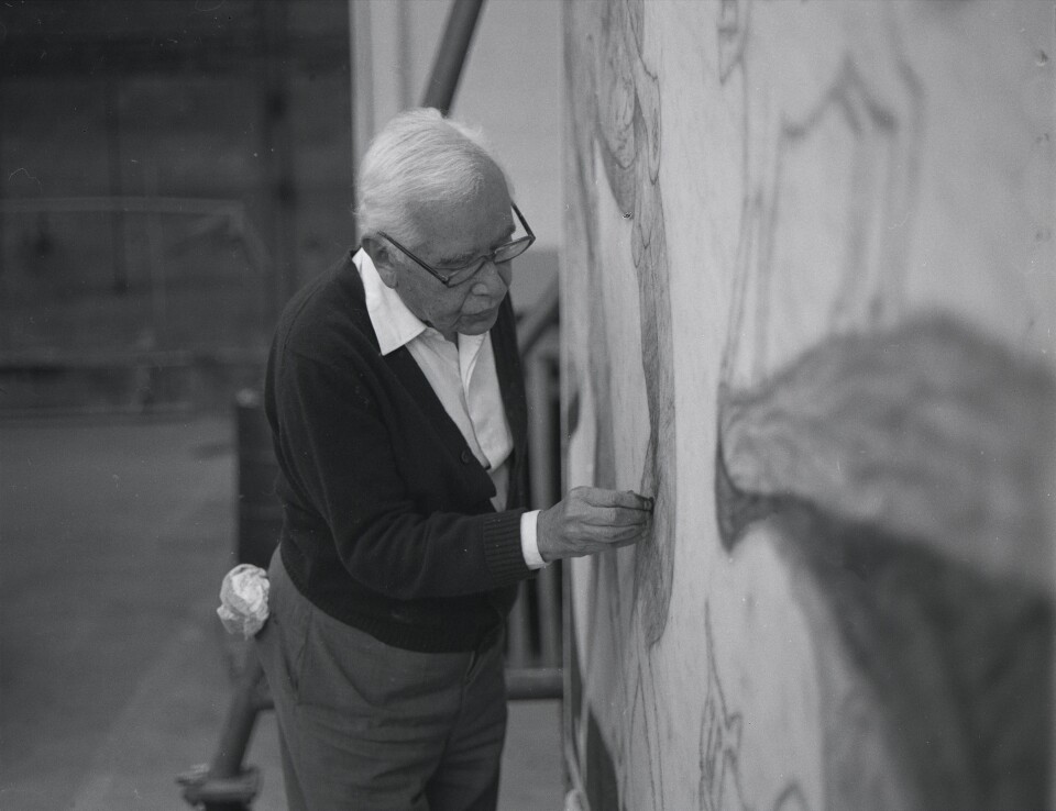 A Hispanic man with white hair and wearing glasses, a white dress shirt, a dark colored coat and light grey slacks, is drawing an object on a stone wall