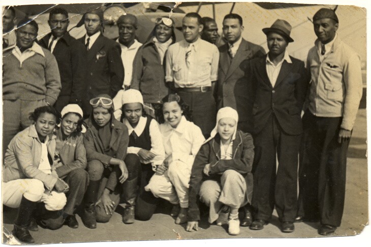16 men and women either crouch or stand in front of an airplane for a group photo