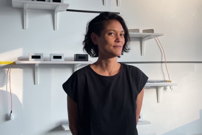 Lauren Lee McCarthy wearing a black top with her hands behind her back standing in front of a white wall.