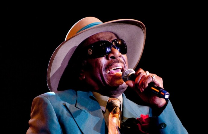 A Black man in a blue suit wears a wide-brim gat as he sings into a mic. He has sunglasses on.