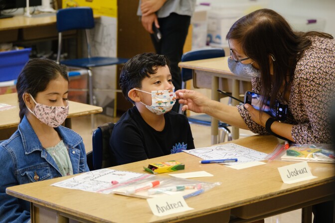 A principal reaches toward a child who's seated at a desk wearing a facemask that's not currently covering his nose.