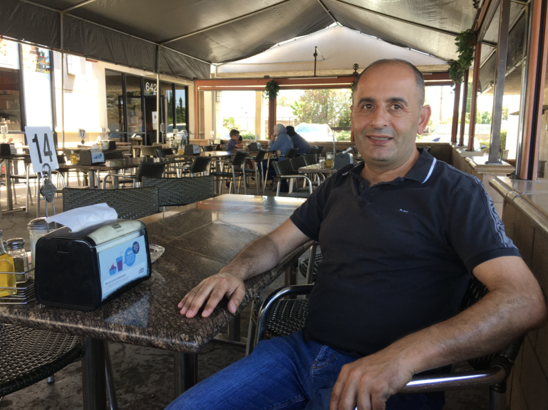 A man in a dark polo shirt and jeans seated in the shade of a restaurant patio.