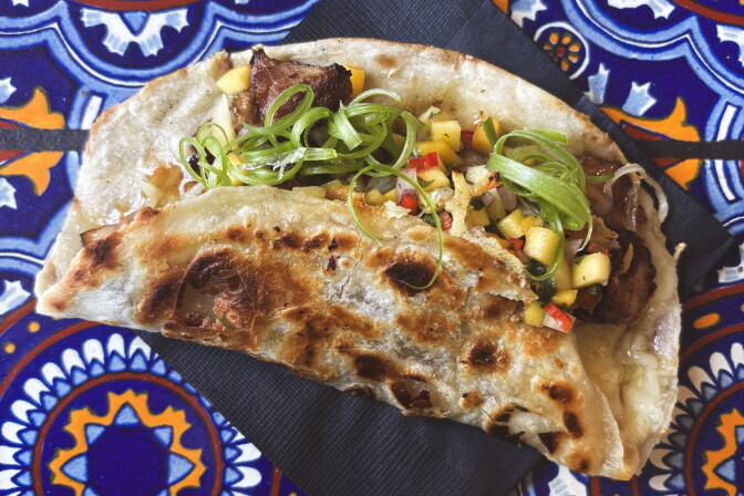 A folded flour tortilla taco filled with jerk chicken, mango salsa, and curled scallions, served on a blue and orange Talavera-tiled table.