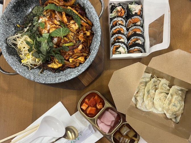 A grey stone pot with caramel-colored squid and onions alongside green perilla leaves and yellow bean sprouts. A container open with tightly rolled kimbap and a cardboard box opened to reveal neatly shaped dumplings. 