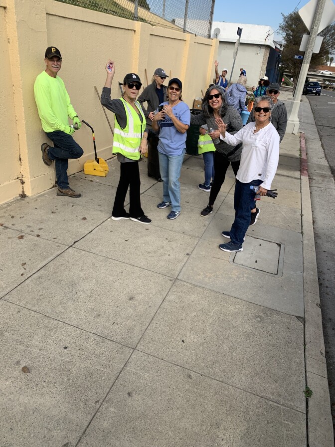 A group of volunteers smiling and waving on a city sidewalk. Several are wearing neon green vests, hats and sunglasses. 
