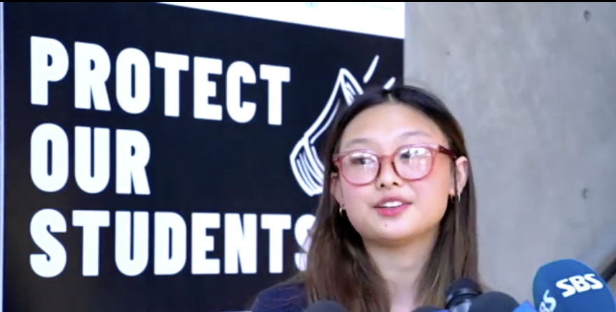 A young Asian woman wearing red glasses with brown hair and a blue shirt stands in behind dark microphones and in front of a black banner that reads "Protect Our Students" in white lettering