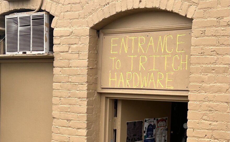 The top two-thirds of a door and entry way to a brick building, all painted the same shade of tan. The door is propped open, and the words "Entrance to Tritch Hardware" has been scratched into the paint above the doorway, revealing a bright yellow below.