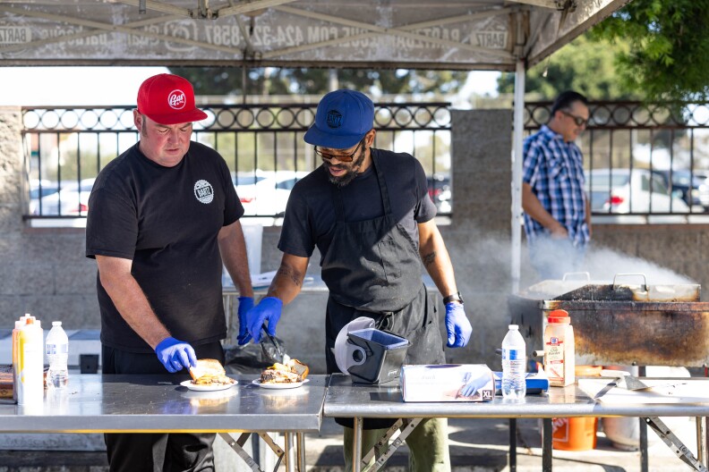 Two men stand behind a metal table beneath a pop-up tend, preparing smashburgers for customers. Smoke is wafting up off a hot grill in the background.