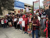 A group of dozens of striking mental health workers and their supporters stand at a rally in Pasadena. They mostly wear red colors and hold protest signs against Kaiser. 