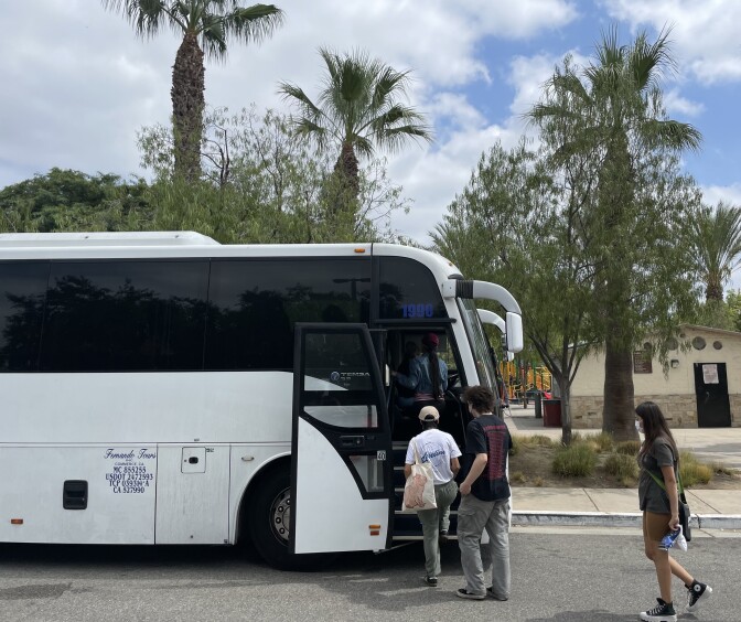 Three people make their way onto a large, commercial. A park restroom is in the background, along with palm trees and a cloudy sky. 