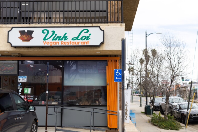 A corner restaurant in a strip mall has a white, green and orange sign saying Vinh Loi above a glass exterior which shows the inside of the dining room. A grey car is parking in front of the building