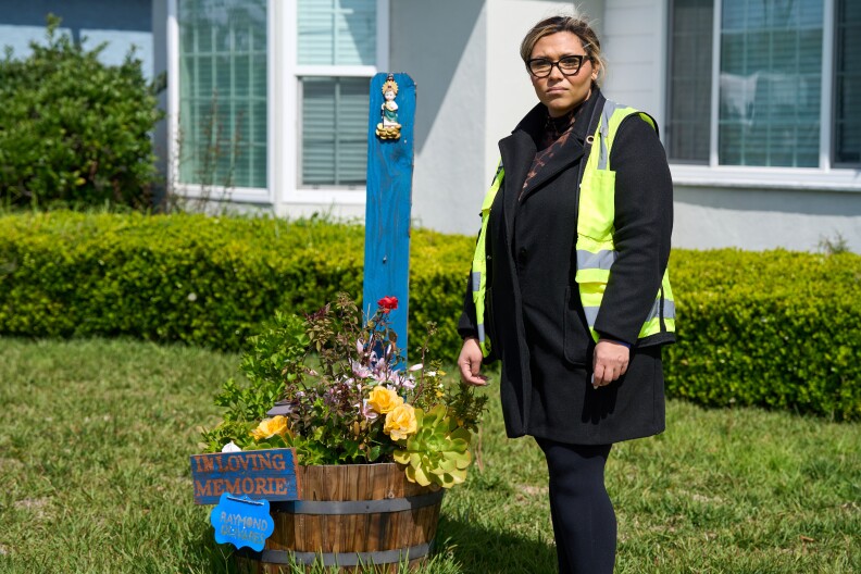 A woman wearing a neon green work jacket and glasses stands next to a memorial consisting of a bucket of flowers and a wooden sign that reads "In Loving Memorie" resting next to a blue wooden post.