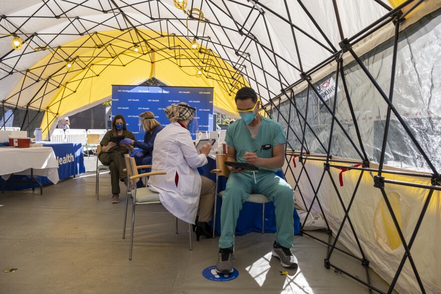 A pop-up tent covers an area where masked health care workers are seated in scrubs and white coats.