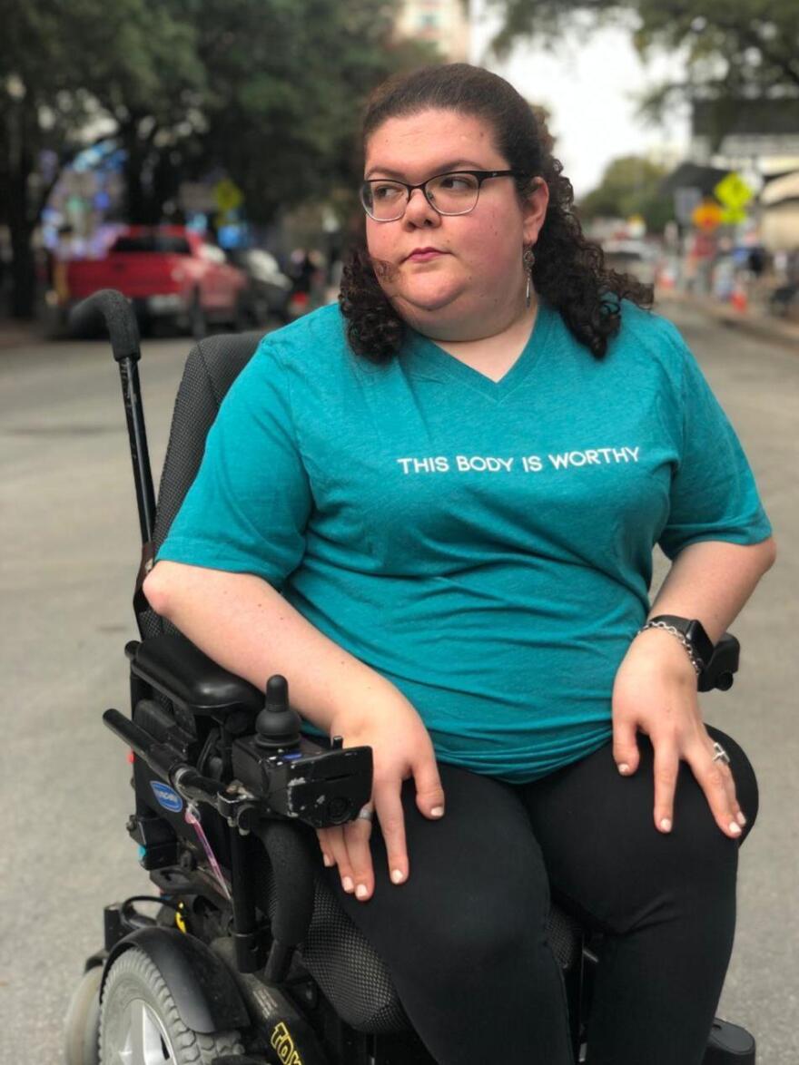Emily Ladau, a white woman, sitting in a wheelchair and wearing glasses and a blue shirt with the text "My Body is Worthy."