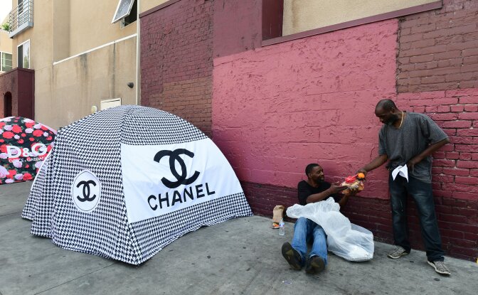 A man offers food to a homeless person seated beside designer tent covers covering tents for the homeless on May 30, 2019 in Los Angeles, California, where figures released on June 4, 2019 show the homeless population in Los Angeles County jumped 12 percent over the past year to nearly 59,000 people, including the elderly, young people and families. (Photo by Frederic J. BROWN / AFP)        (Photo credit should read FREDERIC J. BROWN/AFP/Getty Images)