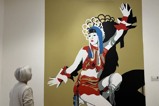 A white hair-bobbed woman in a white sweater looks at large mural of Anna May Wong covering the length of wall. 