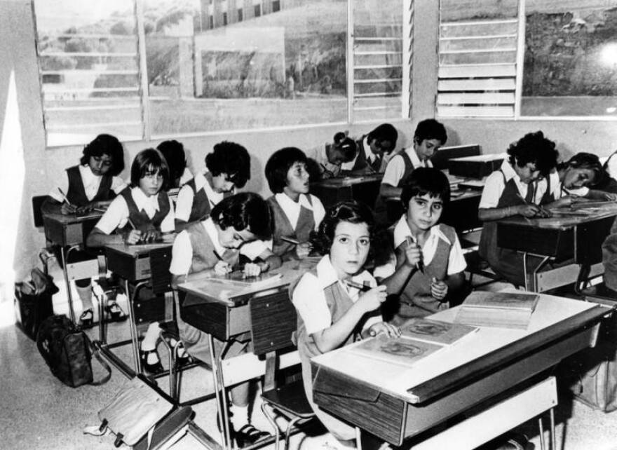 A black and white photo of four rows of children in school sitting at the desks in pairs. They are all wearing uniforms, and there is a window behind them through which you can see the outside. The two children in the front row are looking up at the camera.