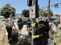 Volunteers gather their trash bags after combing the sidewalks along a Central Avenue intersection on a bright day. One worker has on a white tank-top and black pants, and the other has a black shirt. Both are wearing gloves.