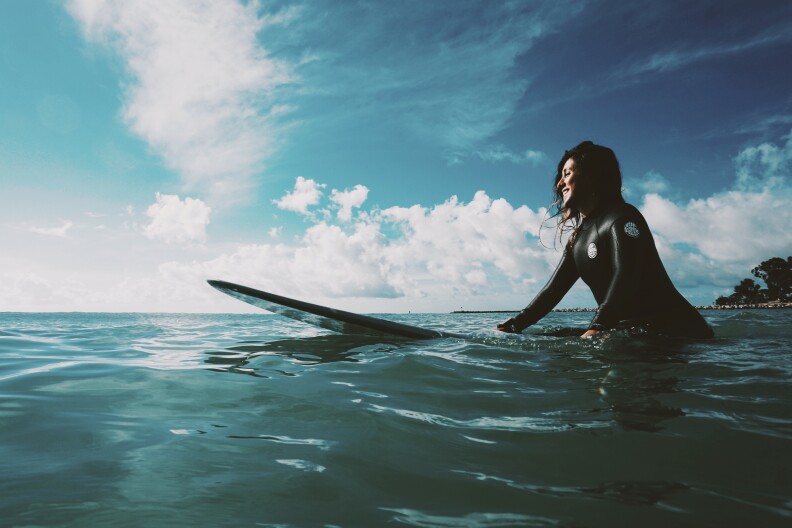 A smiling woman rests on a surf board in the middle of the ocean.