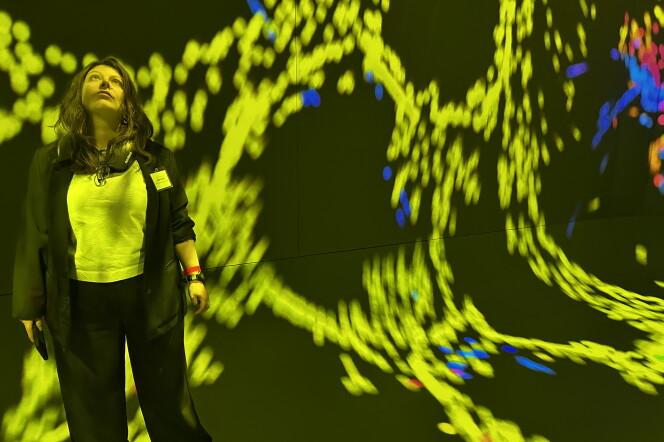 A woman standing in front of an art exhibition comprised of hundreds of yellow-green dots, interspersed with occasional blue dots.