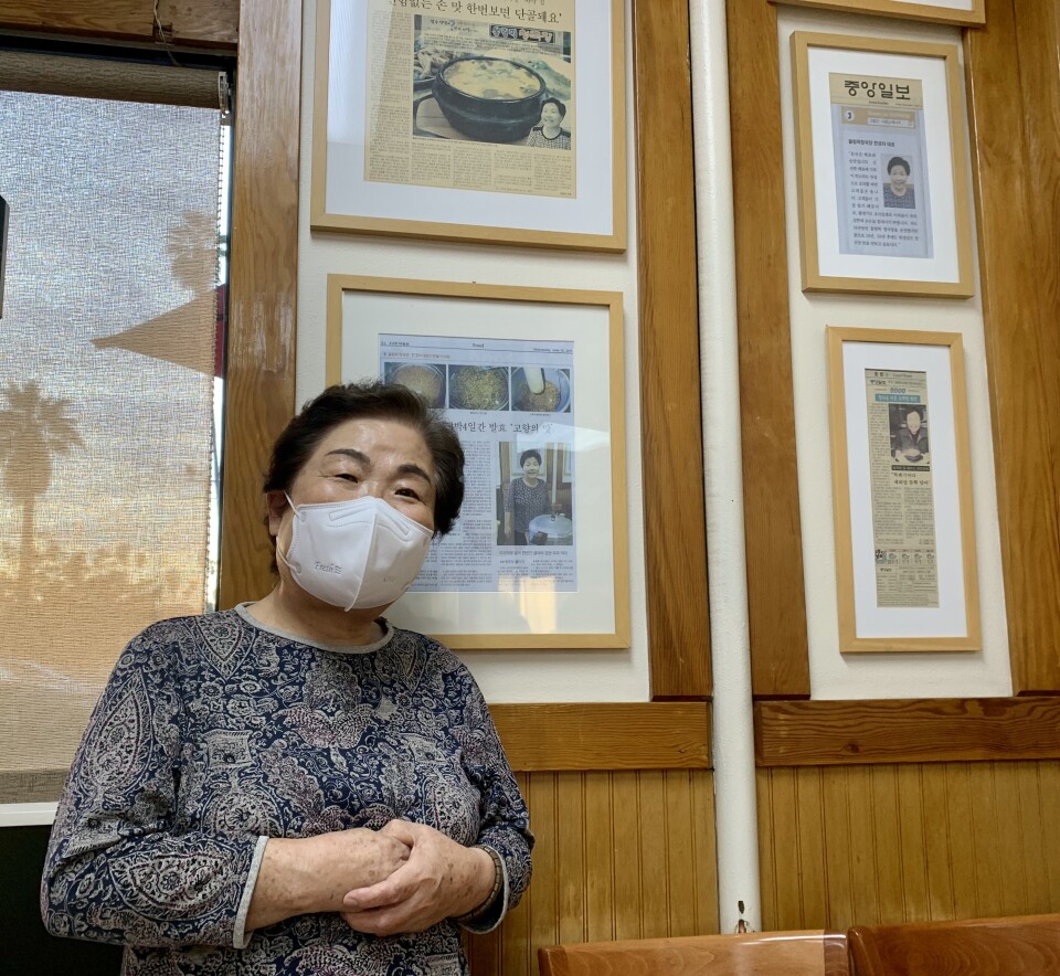 Kyong Han stands next to framed write-ups in Korean-language papers about the Olympic Restaurant. A sunset peeks through a window over her shoulder. 