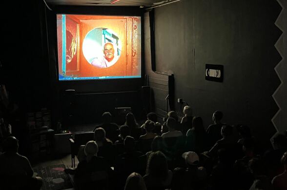 A crowd watches a film projected onto a screen in a dark room. The screen features a man peering out to the audience through an orange circular window.