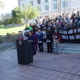 A person speaks at a podium at the steps of L.A. City Hall as a crowd behind her holds banners. One reads"Clean Air & Health Over Big Oil Wealth."