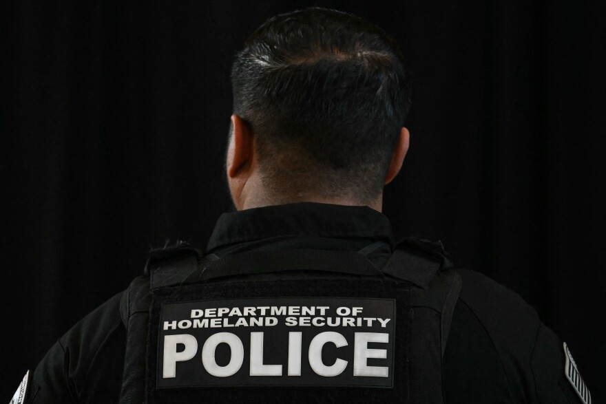 A man with his back to the camera is wearing a black vest that reads in white text "DEPARTMENT OF HOMELAND SECURITY POLICE"