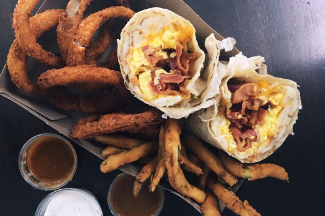 onion rings, breaded and fried zucchini sticks and a breakfast burrito filled with eggs, bacon and cheese sit in a brown cardboard box