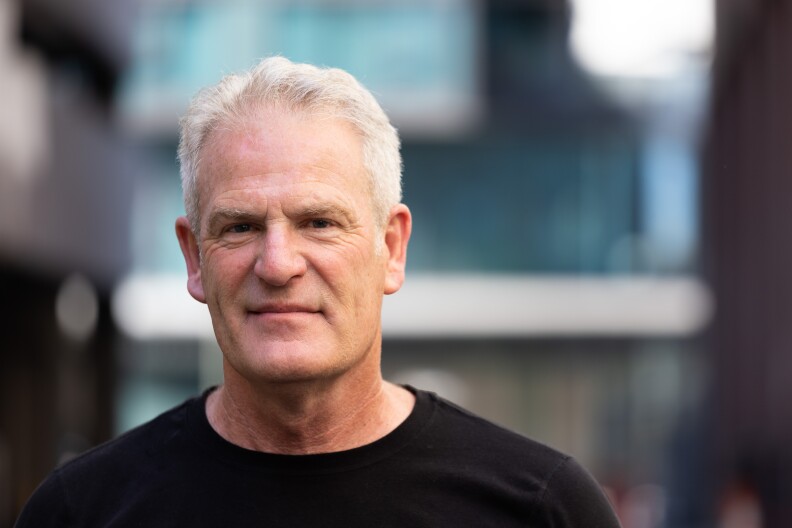 A close up portrait of an older white man with short white hair wearing a black t-shirt. The background is out of focus.
