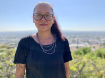A 40-year-old Cambodian American woman in glasses wearing a black shirt and three beaded necklace strands faintly smiles as she stands in a park with trees and waterfront in the background. 