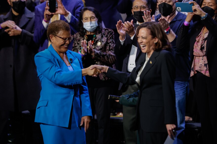 Karen Bass, a light-skinned Black woman wearing a blue pantsuit, shakes hands with Vice President Kamala Harris, a light-skinned Black woman in a black pantsuit. Several people stand behind them clapping.