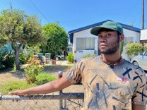 Tryron Ramsey stands in front of his grandfather's house with his right arm resting on the fence. He's wearing a green hat, a necklace he says he made himself, and a graphic t-shirt.