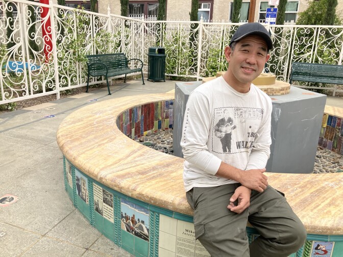 A man in a baseball cap, white graphic t-shirt and olive green pants sits at a fountain that has a yellow stone top and blue, colorful tiles depicting various images.