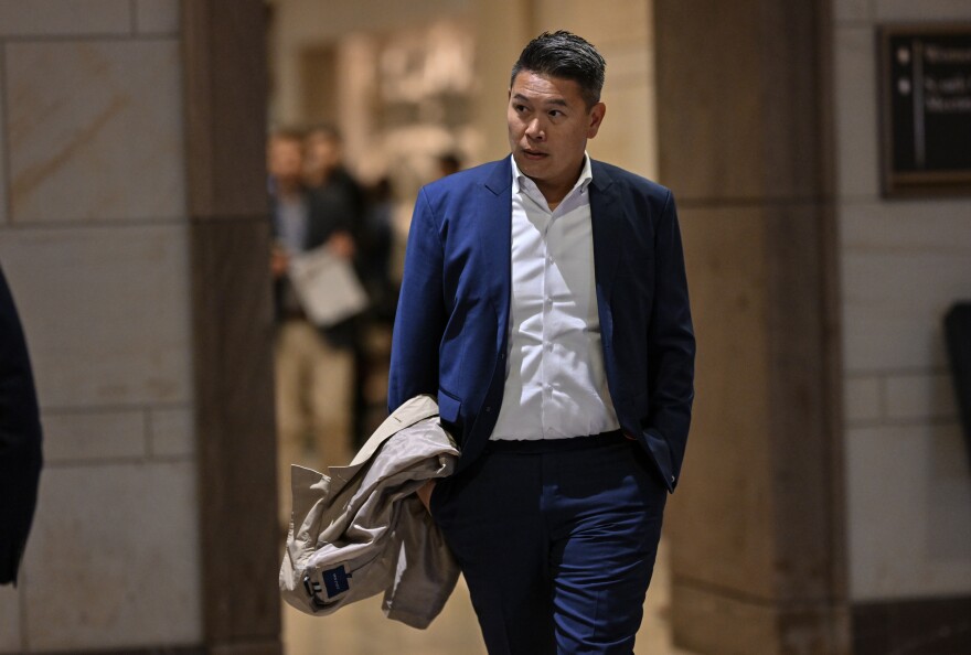 A Vietnamese American man in his early 40s, wearing a royal blue suit and white shirt, walks through a white walled room at the U.S. Capitol, carrying a beige jacket.