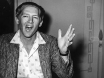 A black and white photo of Jerry Lee Lewis, a then-20-something white man, with his mouth open as if singing or speaking and his right hand gesticulating in the air. 