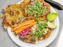 Overhead view of a white paper plate holding three tacos on crisp, griddled tortillas: one folded with melted cheese along the edges, one topped with mushrooms, refried beans, red onion, and cilantro, and one topped with grilled chicken, onion, and cilantro. Carrot sticks, pickled red onions, and lime wedges sit alongside.