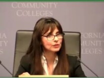 A woman seated with brown hair and glasses speaks at a conference.