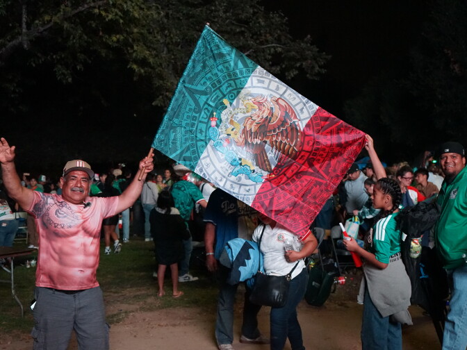 A medium skin toned man holds a large Mexican flag, his arms raised. It's nighttime. A young girl wearing a green Mexico jersey helps him hold the flag.