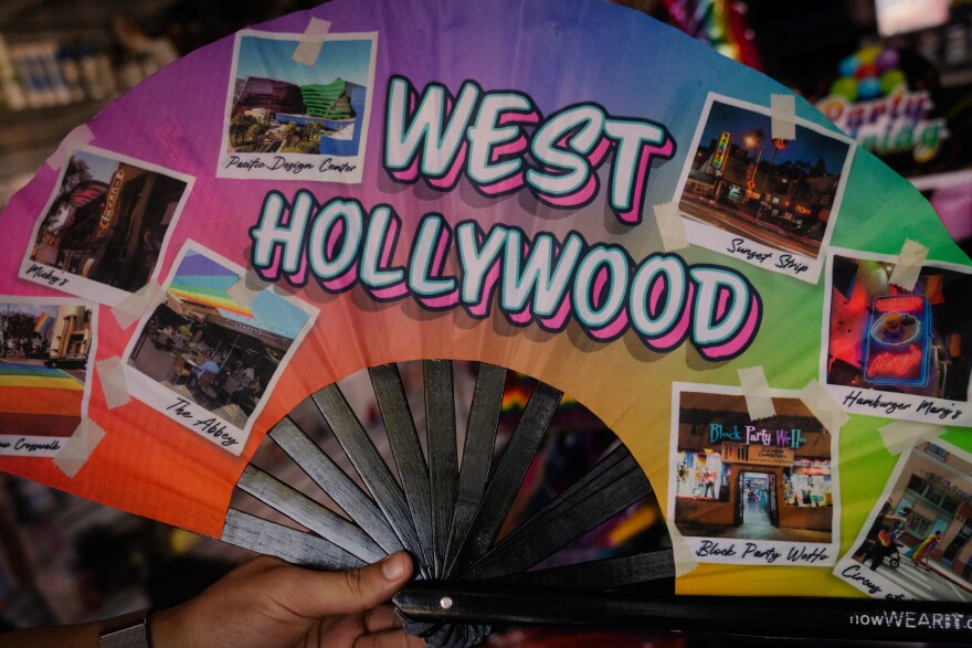 A close up of a person holding a hand fan with a rainbow gradient. It has big letters that say West Hollywood on it and pictures of iconic places, like the Abbey, Hamburger Mary's, Block Party WeHo, and Mickey's